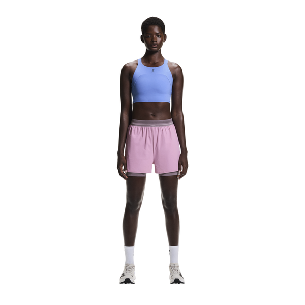 En person står og kigger fremad iført en lyseblå sports-bh, On 3" Performance 2/1 Shorts til kvinder i Sakura/Dewberry, hvide sokker og hvide sneakers på en almindelig hvid baggrund.
