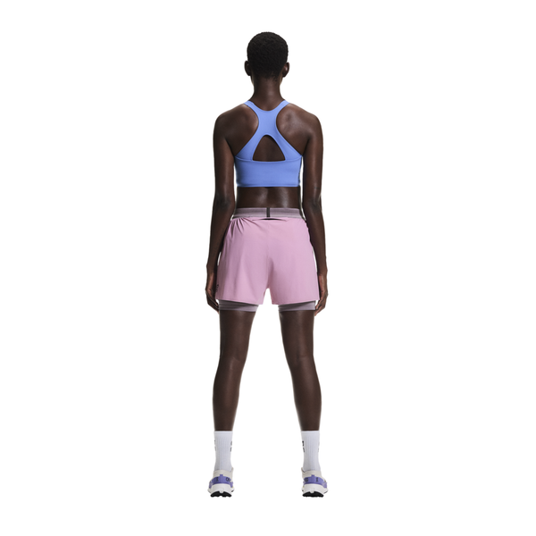 En person står med front mod kameraet iført en lyseblå sports-bh, On 3" Performance 2/1 Shorts for dame i Sakura/Dewberry, hvide lægstrømper og hvide sneakers på en almindelig hvid baggrund.