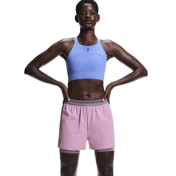 En person står med hænderne på hofterne iført en lyseblå atletisk crop top og On 3" Performance 2/1 Shorts til kvinder i Sakura/Dewberry med grå linning. Baggrunden er helt hvid.