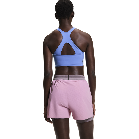 En person med kort hår ses bagfra, iført en lyseblå racerback sports-bh og On 3" Performance 2/1 Shorts Dame i Sakura/Dewberry, stående mod en almindelig hvid baggrund.