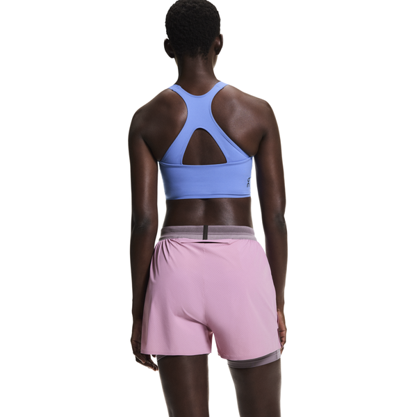 En person med kort hår ses bagfra, iført en lyseblå racerback sports-bh og On 3" Performance 2/1 Shorts Dame i Sakura/Dewberry, stående mod en almindelig hvid baggrund.