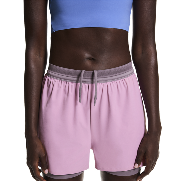 En person iført blå crop top og On 3" Performance 2/1 Shorts, Dame, i Sakura/Dewberry med grå linning og snøre, står med armene ned langs siden. Billedet er beskåret for at vise overkroppen og overbenene.