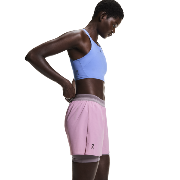 En person iført en lyseblå sports-bh og On 3" Performance 2/1 Shorts, Dame, Sakura/Dewberry, står i profil mod en hvid baggrund og justerer shortsens grå linning.