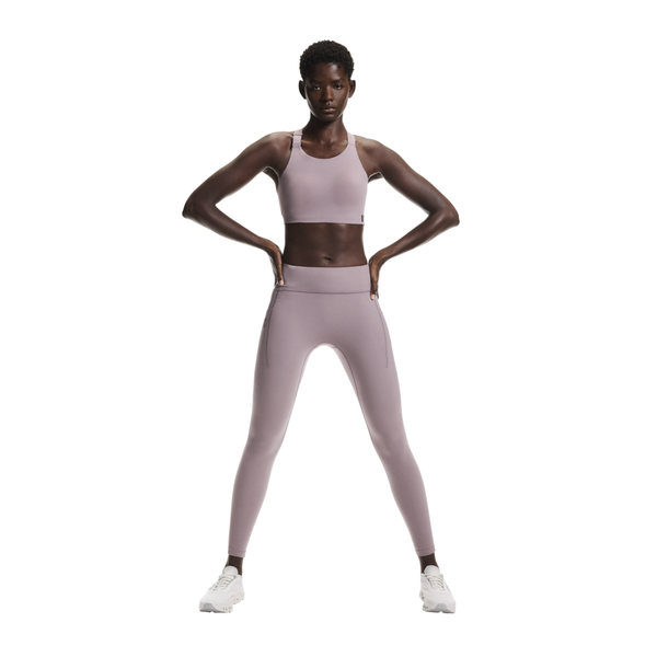 En person står med hænderne på hofterne iført lilla On Performance Tights, Dame, Heron og en matchende sports-bh med hvide sneakers på en almindelig hvid baggrund.