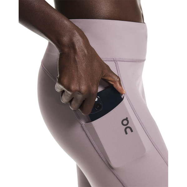 En person lægger en smartphone i sidelommen på On Performance Tights, Dame, Heron - disse løbetights er lavet af DryTec-materiale for komfort og åndbarhed og har On-mærkets logo.