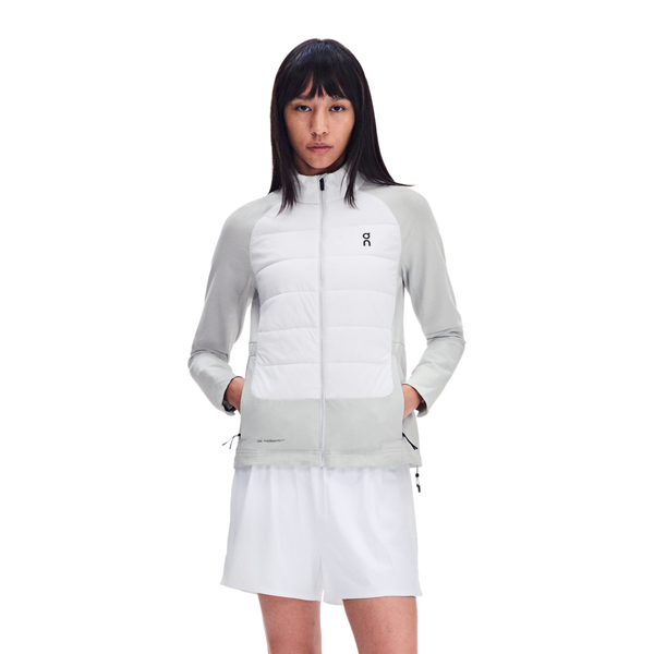En person iført en On Weather Hybrid-jakke, Dame, Glacier/Frost, med et hvidt og lysegråt, vind- og vandafvisende design med et quiltet forstykke, kombineret med hvide shorts.