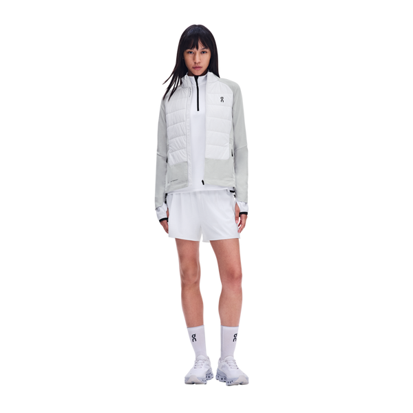 En kvinde har en hvid og lysegrå On Weather Hybrid Jacket, Dame, Glacier/Frost på over en sports-top og shorts, og looket fuldendes med matchende hvide sneakers og sokker.