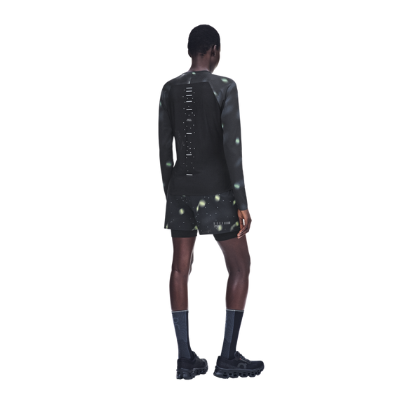 En person set bagfra, iført en On Performance Long-T Lumos, Dame, sort, med sorte shorts med et reflekterende, plettet mønster, sorte sokker og sorte sportssko.