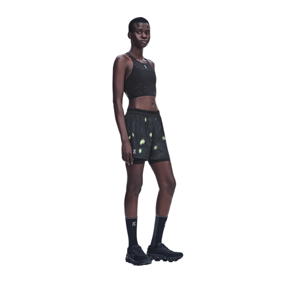 En kvinde iført en sort, kort sports-top, On Performance Shorts Lumos, Dame, sort, sorte sokker og sorte løbesko.