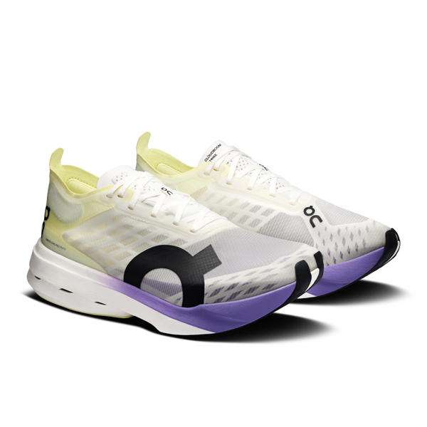 Et par On Cloudboom Strike løbesko til damer i Juniper/Limelight med gennemsigtig mesh-overdel, sorte logoer, lysegrønt inderfor og tykke hvide såler med en lilla accent foran.