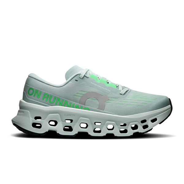 En ON Cloudmonster 3 løbesko til kvinder i farven Ivory/Topaz har en overdel i mesh, grå snørebånd, grøn ON RUNNING-tekst og en tyk hvid sål med CloudTec-støddæmpning og udskårne detaljer. Vist på en ensfarvet hvid baggrund.
