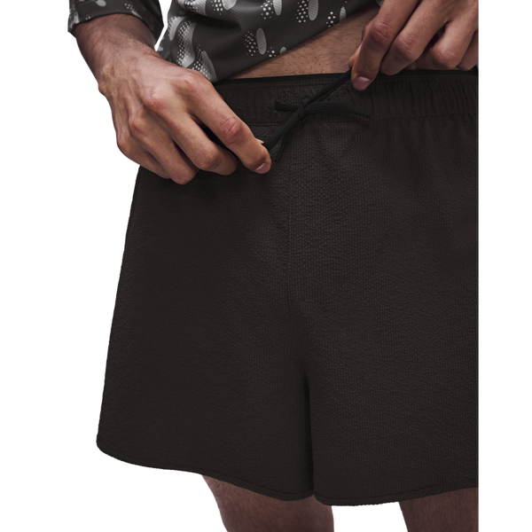 En person strammer snoren i et par ON Performance Volt Shorts, Herre, Espresso. Personen har en mønstret skjorte på med opsmøgede ærmer, og på det beskårne billede ses kun overkroppen og de øverste dele af benene.