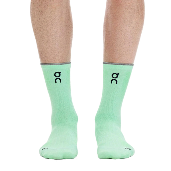 En person iført ON Elite Run Sock High Hyper-strømper i farven Celeste/Verdite, med et sort ON-logo og et tyndt gråt bånd øverst, står på en hvid baggrund. Disse unisex tekniske løbestrømper, der er designet til løbetræning, viser kun underbenene og fødderne på billedet.