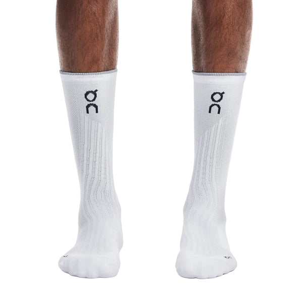 En person står på en hvid baggrund og har et par hvide On Elite Run Sock High Hyper på. Disse unisex-løbestrømper i høj kvalitet har ribdetaljer og et sort On-logo. Personens underben er synlige, hvilket understreger, at strømperne er velegnede til løb.