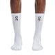 En person står på en hvid baggrund og har et par hvide On Elite Run Sock High Hyper på. Disse unisex-løbestrømper i høj kvalitet har ribdetaljer og et sort On-logo. Personens underben er synlige, hvilket understreger, at strømperne er velegnede til løb.