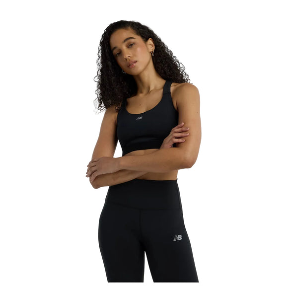 En kvinde bærer New Balance Nb Essential Train Bra i sort med korslagte arme og selvtillid mod en hvid baggrund - perfekt klædt på til enhver træningssession.