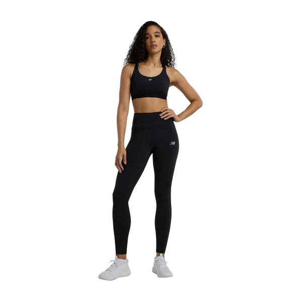 En kvinde poserer på en hvid baggrund iført New Balance Nb Essential Train Bra til kvinder i sort, sammen med matchende leggings og hvide sneakers. Hun har langt krøllet hår og en hånd på hoften, klar til at træne.