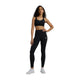 En kvinde poserer på en hvid baggrund iført New Balance Nb Essential Train Bra til kvinder i sort, sammen med matchende leggings og hvide sneakers. Hun har langt krøllet hår og en hånd på hoften, klar til at træne.