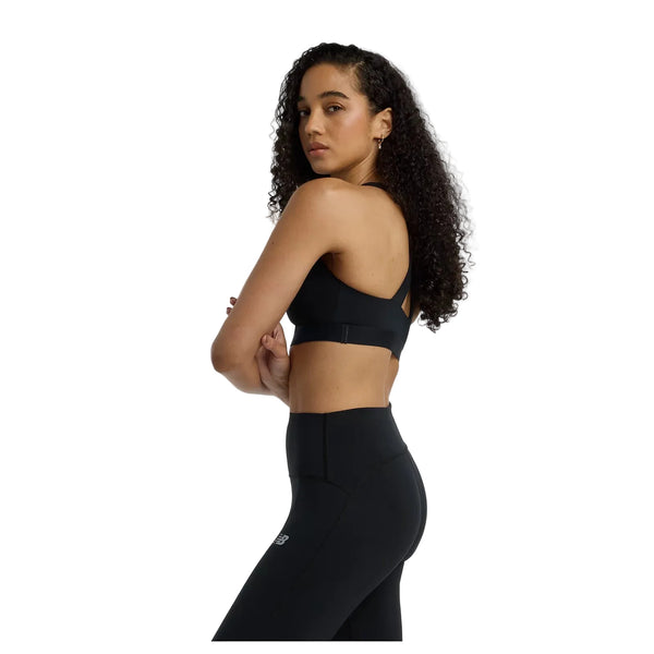 En kvinde med langt krøllet hår modellerer New Balance Nb Essential Train Bra, Dame, Black, stående sidelæns med korslagte arme og kigger sig over skulderen på en almindelig hvid baggrund.