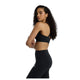 En kvinde med langt krøllet hår modellerer New Balance Nb Essential Train Bra, Dame, Black, stående sidelæns med korslagte arme og kigger sig over skulderen på en almindelig hvid baggrund.