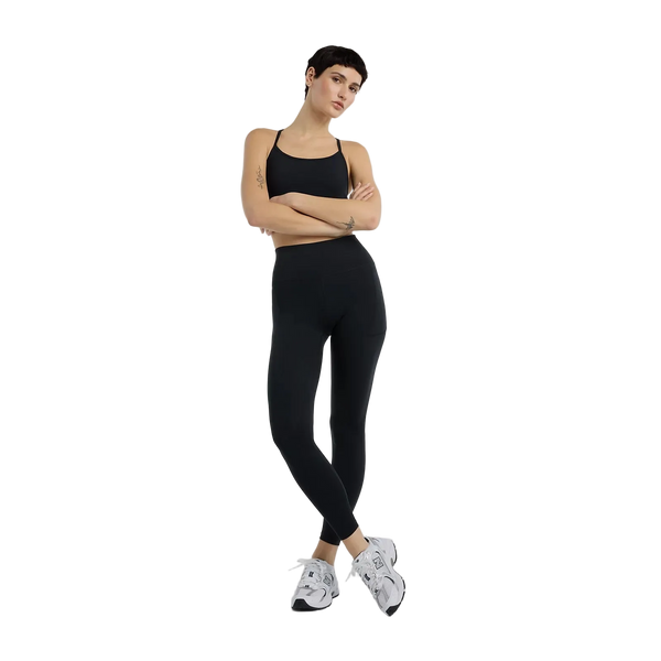 En kvinde med kort mørkt hår står med armene over kors iført New Balance Essential Yoga Bra, Dame, Black og leggings i blødt, åndbart materiale sammen med hvide sneakers på en almindelig hvid baggrund.