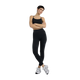 En kvinde med kort mørkt hår står med armene over kors iført New Balance Essential Yoga Bra, Dame, Black og leggings i blødt, åndbart materiale sammen med hvide sneakers på en almindelig hvid baggrund.