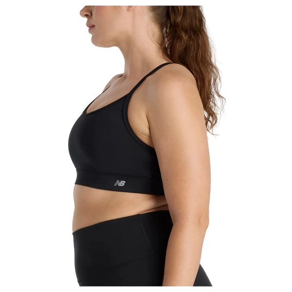 En kvinde med langt brunt hår i hestehale bærer New Balance Essential Yoga Bra, Dame, Black og sorte leggings, vist i profil mod en almindelig hvid baggrund. Bh'en har blødt åndbart materiale og et lille New Balance-logo.