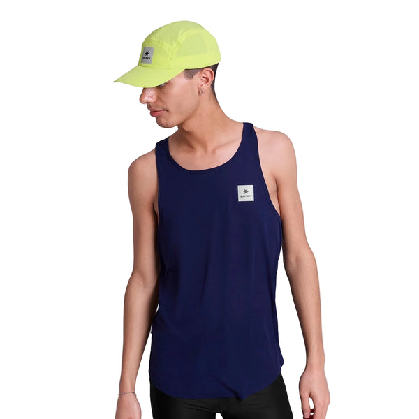 En person står på en hvid baggrund og kigger ned og til siden iført en marineblå hurtigtørrende tanktop med brystpatch og den gule Saysky Flow Cap, begge med mærkets firkantede logo.