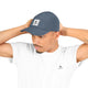En mand iført en grå Saysky Flow Cap og en hvid T-shirt med SAYSKY-logoer står med hænderne bag hovedet foran en ensfarvet baggrund. Unisex-modellen Flow Cap fra Saysky er designet til at være åndbar og behagelig at have på i enhver aktiv livsstil.