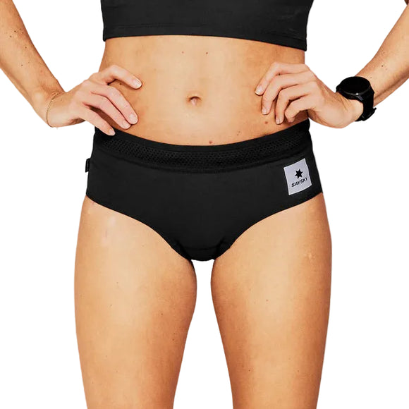 En person iført Saysky Flow+ Race Brief, Dame, Black med et påsyet rektangulært mærke, en sort sportstop og et sort armbåndsur står med hænderne på hofterne mod en hvid baggrund.