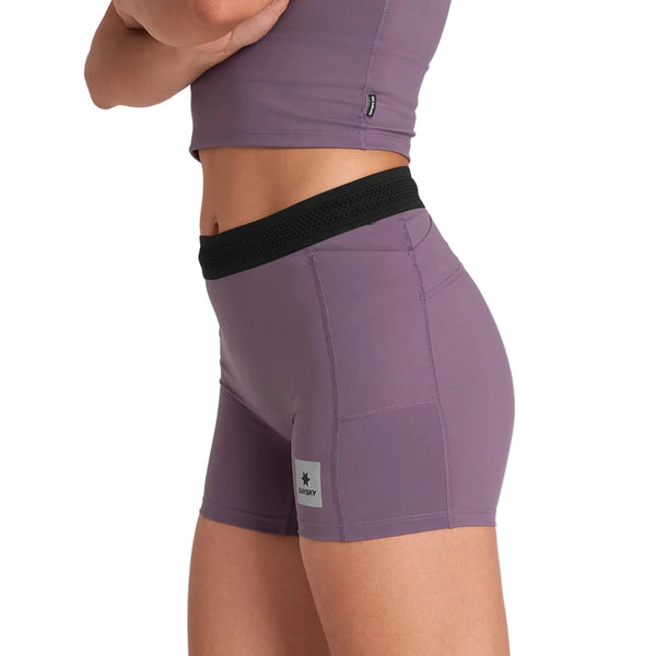 En person i Saysky W Flow+ Race Short Tights 4" til kvinder i lilla, med sort linning og logopatch på venstre ben, parret med en matchende atletisk crop top, står på en almindelig hvid baggrund.