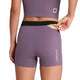 En kvinde i Saysky W Flow+ Race Short Tights 4" i lilla og en matchende top putter en energigel i linningens baglomme. Saysky-logoet er synligt på venstre side af disse stilfulde dame løbetights.