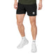 En person står iført sorte Saysky Flow Shorts 5′’ (Herre) med et lille mærke på venstre ben, kombineret med en grøn skjorte, hvide sokker og hvide sportssko. Baggrunden er ensfarvet og lys.