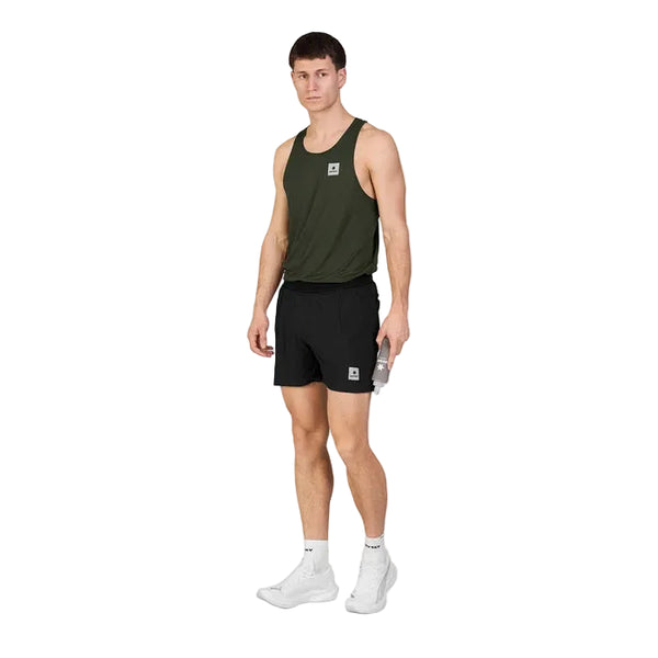 En person iført en mørkegrøn tanktop, Saysky Flow Shorts 5′’ til mænd i sort, hvide sokker og hvide sneakers står på en ensfarvet hvid baggrund og holder en vandflaske.