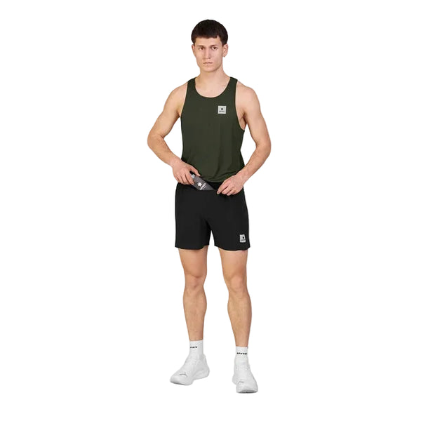 En mand står med ansigtet vendt fremad iført en grøn tanktop, Saysky Flow Shorts 5′’ Herre i sort, hvide sokker og hvide sportssko foran en ensfarvet hvid baggrund. Han holder en sort genstand med begge hænder ved taljen.