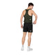 En person iført en grøn tanktop, Saysky Flow Shorts 5′’ Herre i sort, hvide sokker og hvide sneakers står med ryggen til og lægger en genstand i baglommen på sine shorts foran en ensfarvet hvid baggrund.