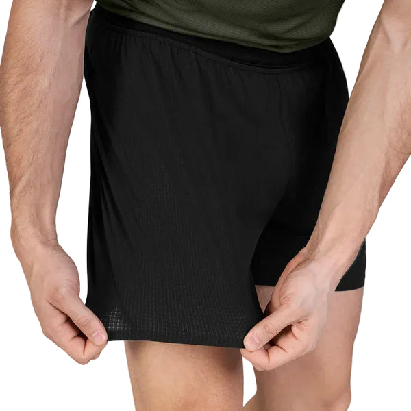 En person iført et par Saysky Flow Shorts 5′’ til mænd i sort og en grøn skjorte står og holder fast i shortsens nederste kant med begge hænder for at vise stoffet frem. Personens arme og en del af lårene er synlige.