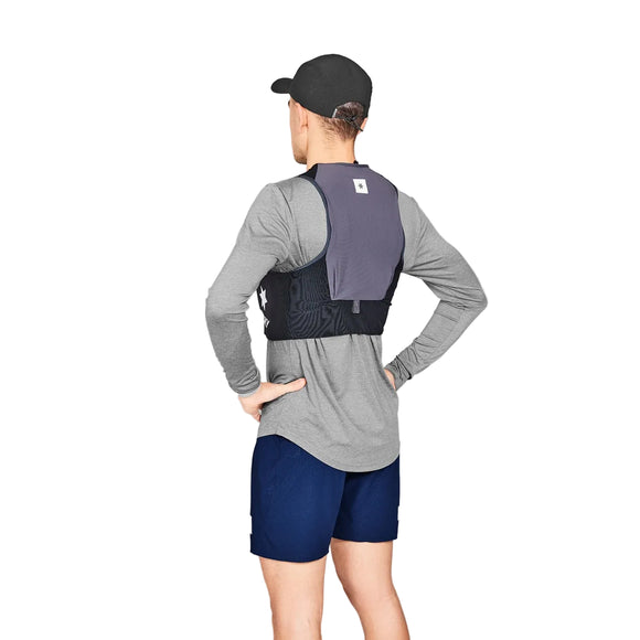 En person står med ansigtet vendt mod kameraet iført en sort Saysky Flow Vest 101 8L (unisex), sort kasket, langærmet grå skjorte og blå shorts med hænderne på hofterne mod en almindelig grå baggrund.