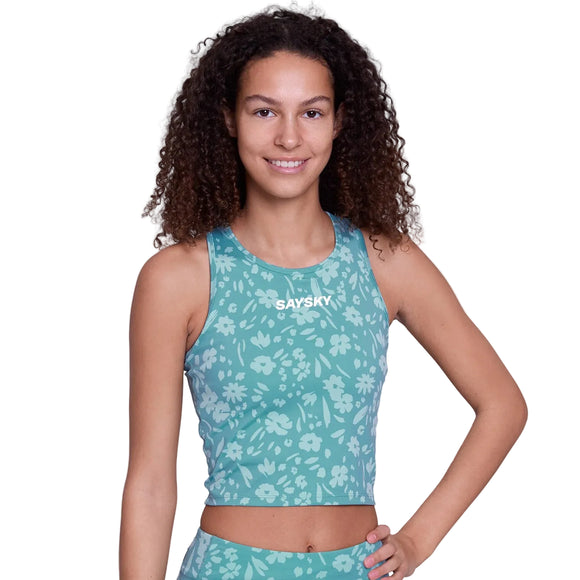 En person med krøllet hår smiler med en hånd på hoften iført Saysky Flower Combat Crop Top, Dame in Light Blue Flower - en ærmeløs model med medium støtte og et hvidt blomstermønster. "SAYSKY" står der på brystet.
