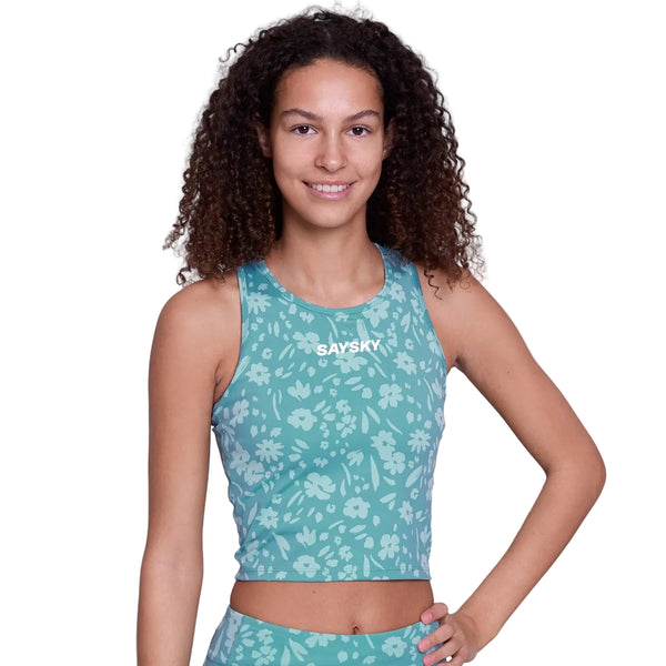 En person med krøllet hår smiler med en hånd på hoften iført Saysky Flower Combat Crop Top, Dame in Light Blue Flower - en ærmeløs model med medium støtte og et hvidt blomstermønster. "SAYSKY" står der på brystet.