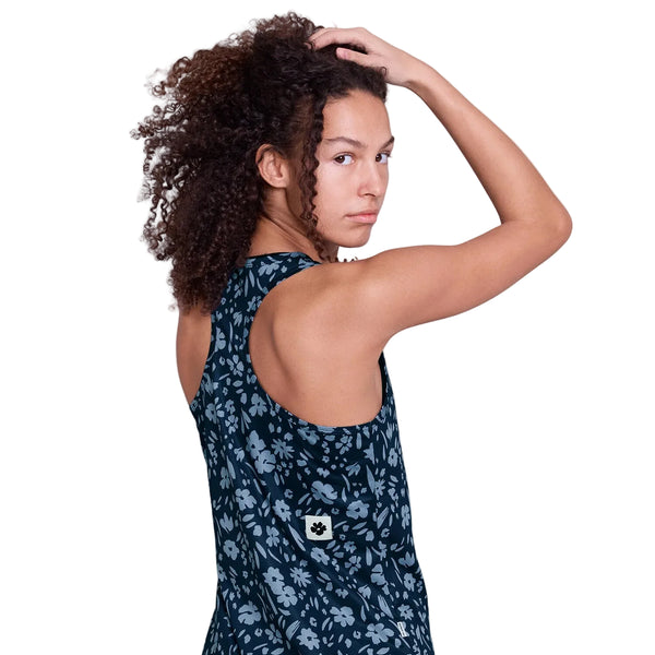 En person med krøllet hår bærer Saysky Flower Combat Singlet, Dame, Blue Flower - mørkeblå med et lyst blomstermønster. Når man kigger sig over skulderen og rører ved hovedet, kan man se et lille hvidt mærke med et poteaftryk på ryggen.