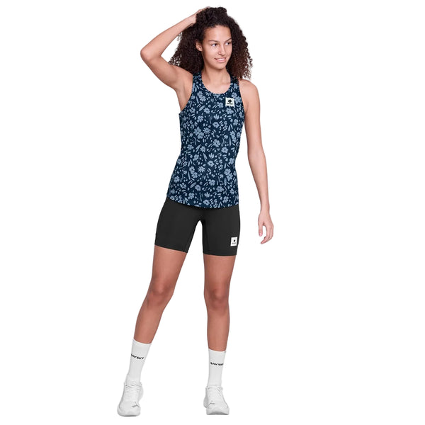 En person står på en hvid baggrund iført Saysky Flower Combat Singlet, Dame, Blue Flower, sammen med sorte shorts, hvide sokker og hvide sneakers. Den ene hånd hviler på hovedet, mens den anden arm hænger ved siden af.