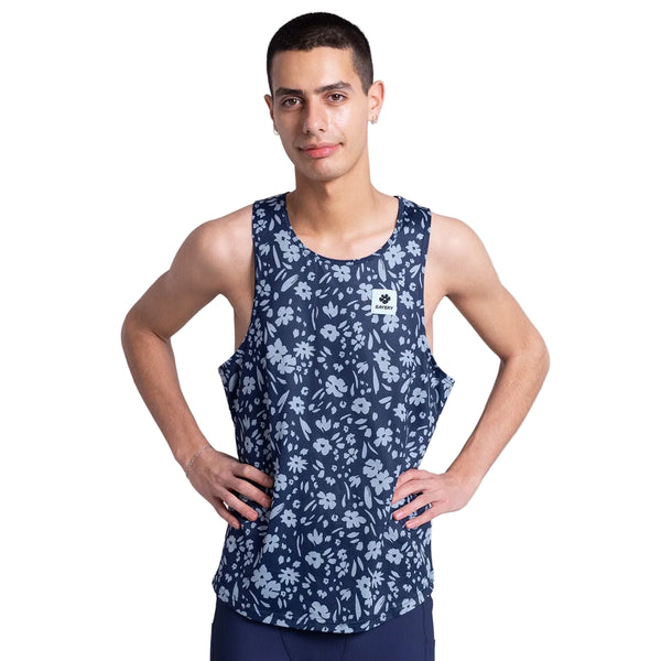 En person står med hænderne på hofterne iført Saysky Flower Combat Singlet, Herre, Blue Flower - en marineblå løbetop med et lyseblåt blomstermønster - og mørke shorts på en almindelig hvid baggrund.