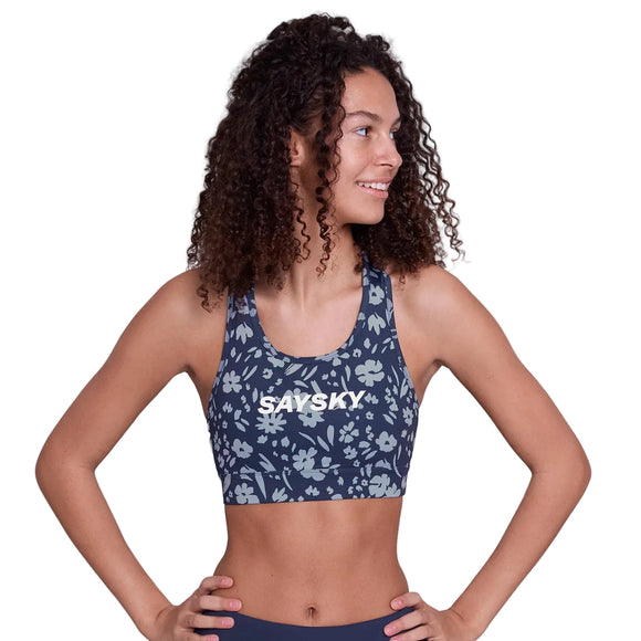 En kvinde med krøllet hår står selvsikkert iført Saysky Flower Combat Sports Bra, Dame, Blue Flower-navy med et lyseblåt blomstermønster og "SAYSKY" på forsiden. Hendes hænder hviler på hofterne, mens hun kigger til siden.