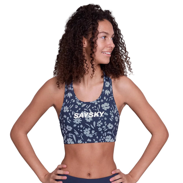 En kvinde med krøllet hår står selvsikkert iført Saysky Flower Combat Sports Bra, Dame, Blue Flower-navy med et lyseblåt blomstermønster og "SAYSKY" på forsiden. Hendes hænder hviler på hofterne, mens hun kigger til siden.