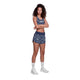 En kvinde med krøllet hår og armene over kors er iført Saysky Flower Combat Sports Bra, Dame, Blue Flower og matchende shorts med hvidt blomstermønster samt hvide sokker og sportssko på en almindelig hvid baggrund.