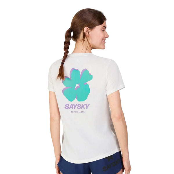 En kvinde med en brun fletning ses bagfra iført en hvid Saysky Flower Combat T-shirt, Dame, med en stor blågrøn og lilla blomstergrafik og "SAYSKY COPENHAGEN" på ryggen. Hun kombinerer den med marineblå shorts.
