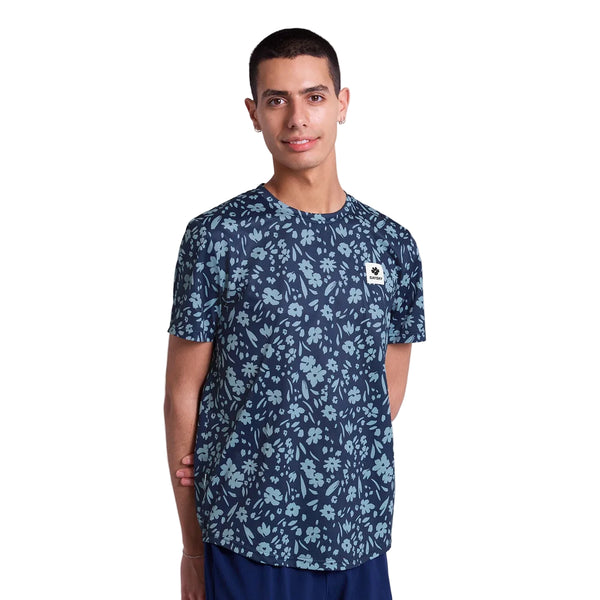 En person står på en hvid baggrund iført Saysky Flower Combat T-Shirt, Herre, Blue Flower, med et lyseblåt blomstermønster og et lille hvidt og gult brystmærke. Armene er afslappede bag ryggen.