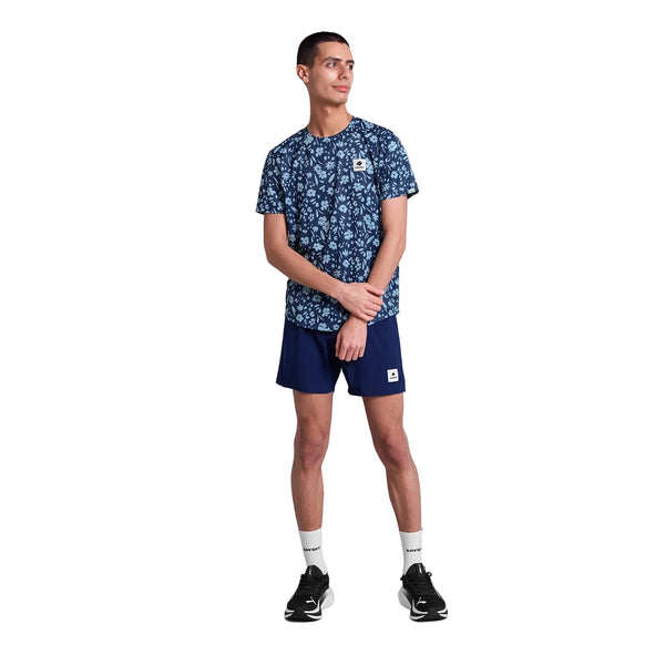 En person iført Saysky Flower Combat T-shirt, Herre, Blue Flower, marineblå shorts, hvide strømper og sorte sportssko står på en hvid baggrund og kigger til siden med hænderne foldet foran sig.