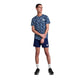 En person iført Saysky Flower Combat T-shirt, Herre, Blue Flower, marineblå shorts, hvide strømper og sorte sportssko står på en hvid baggrund og kigger til siden med hænderne foldet foran sig.